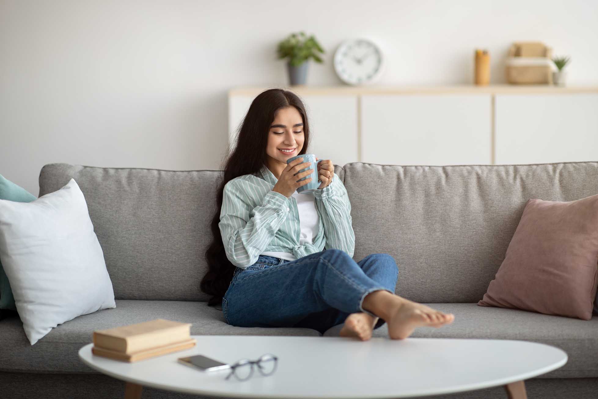 Full length of young Indian woman having coffee break on comfy sofa at home