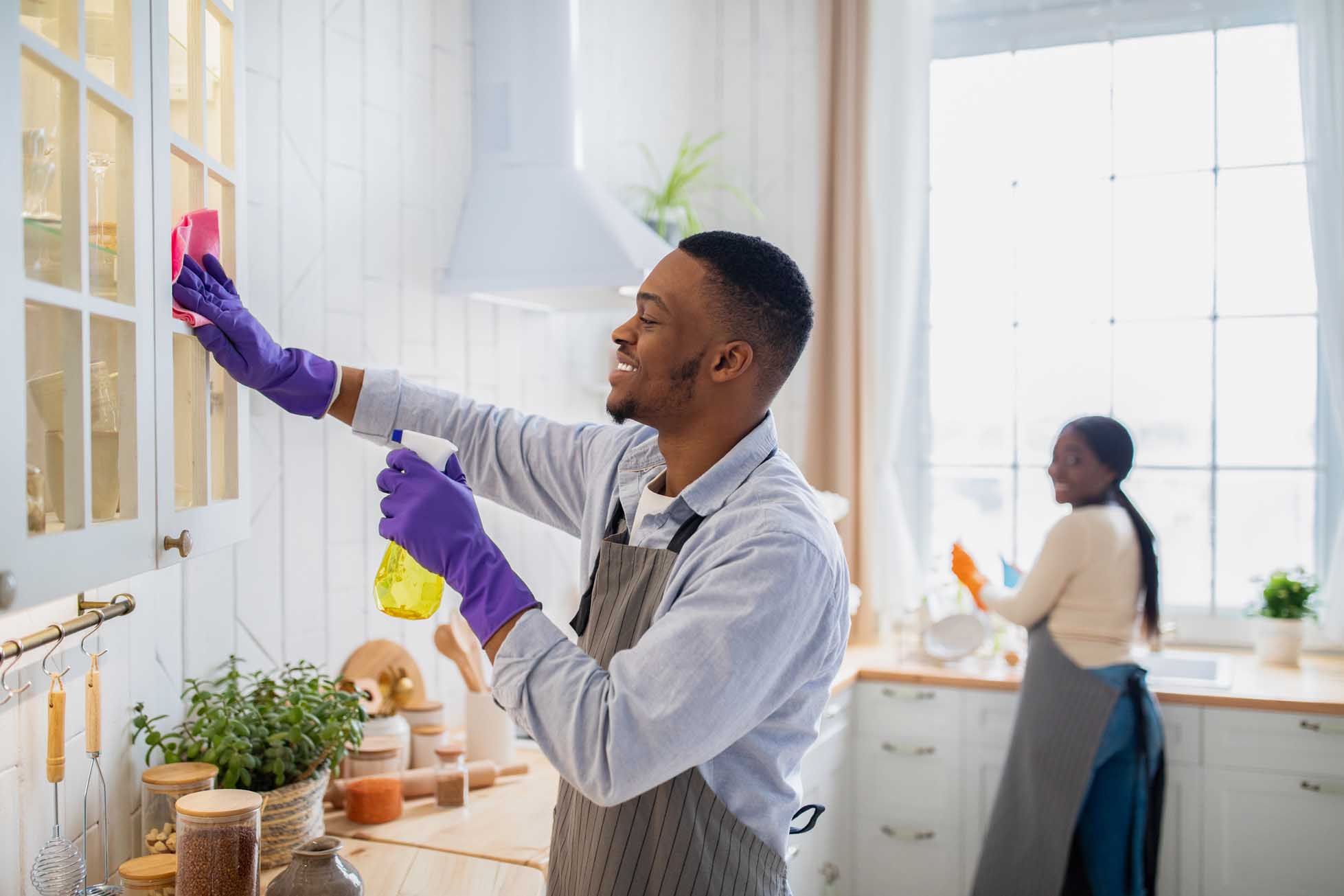 Handsome African American man cleaning kitchen cabinet, his wife washing dishes on background