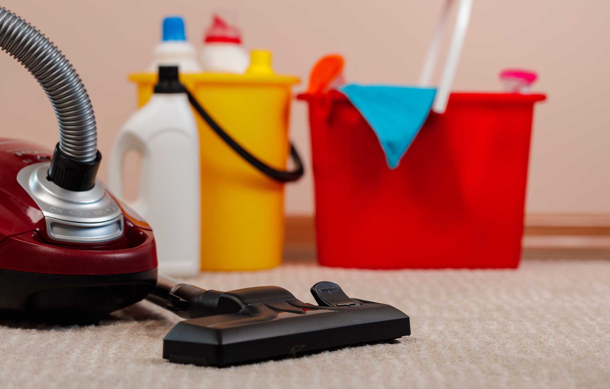 Red vacuum cleaner and plastic bucket with liquid detergents on beige carpet