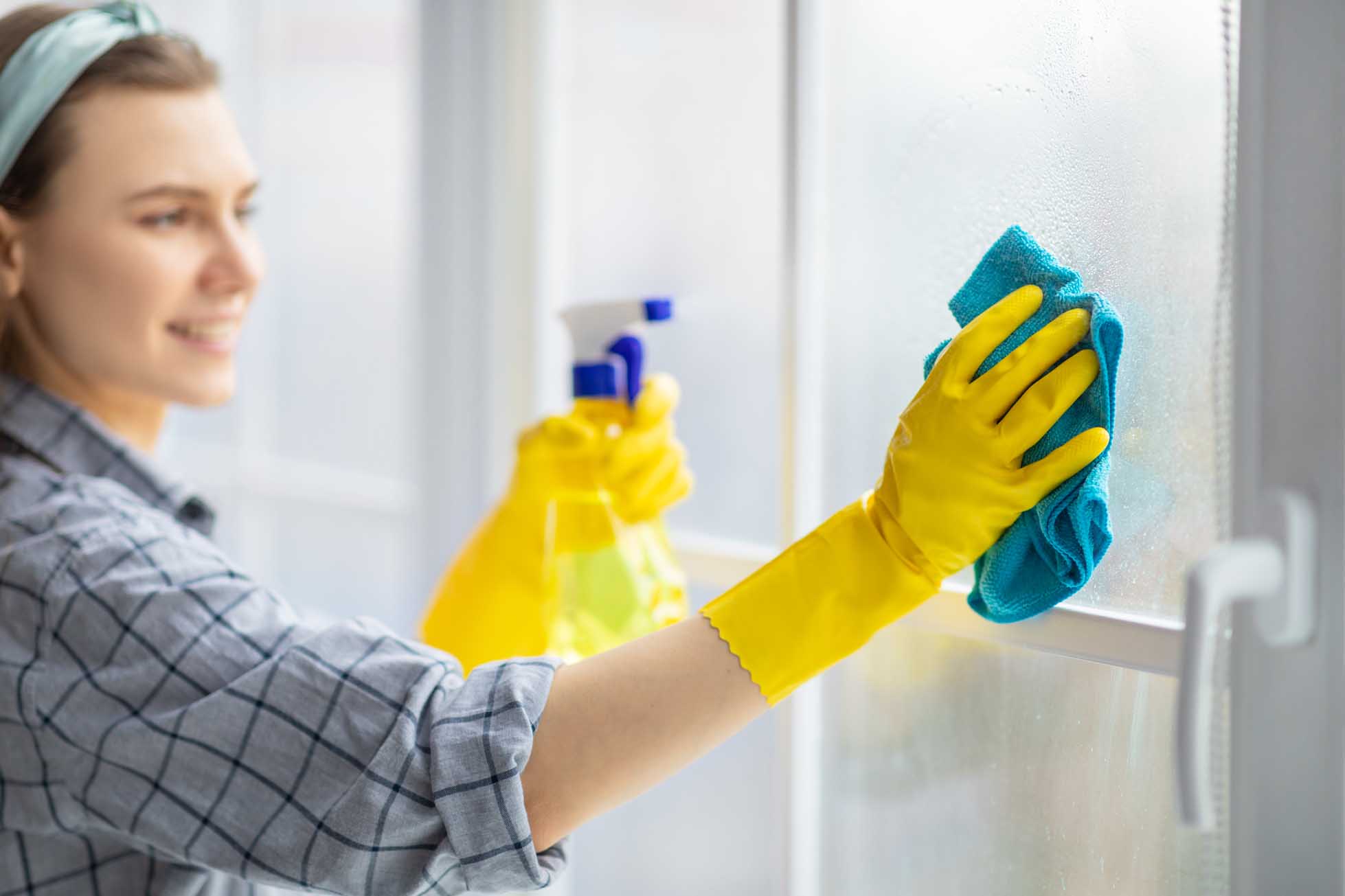 Young housekeeper washing window glass with rag and spray detergent, selective focus